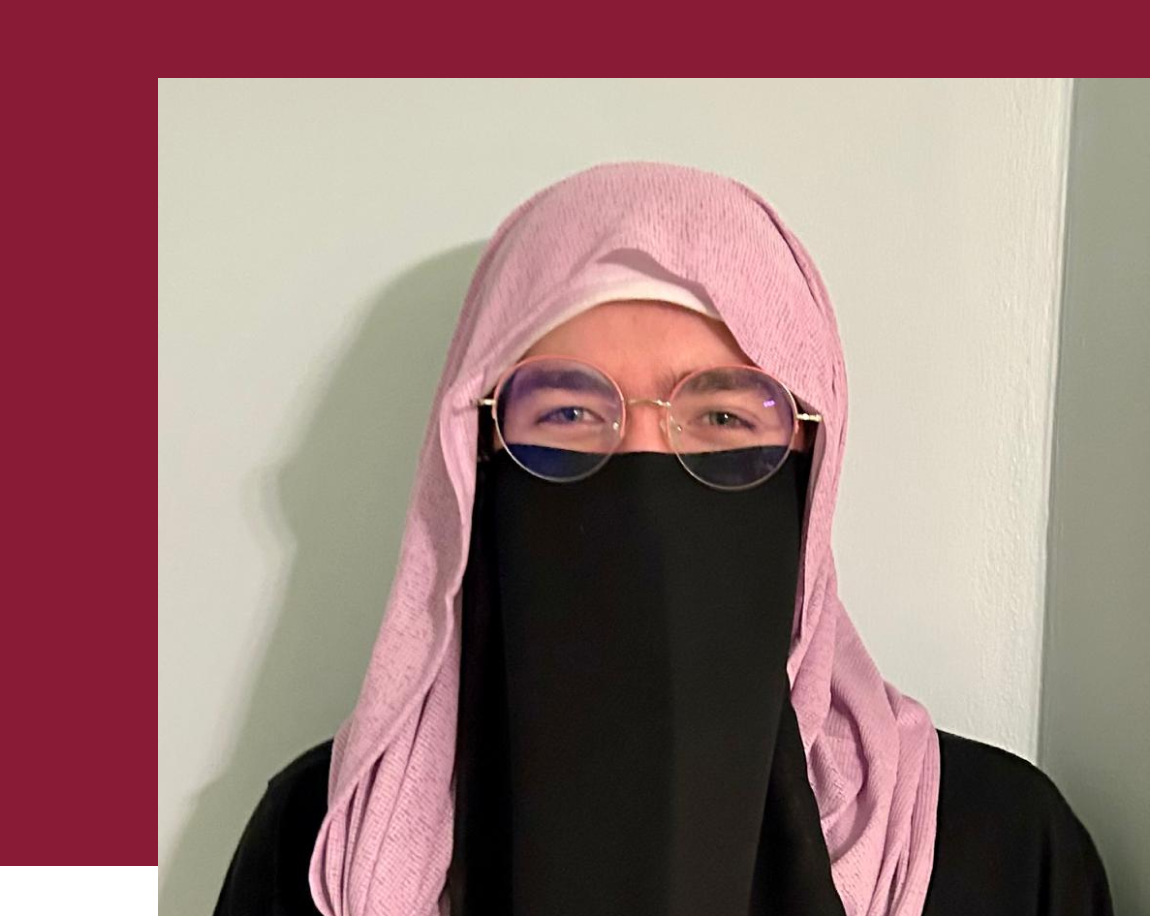 a woman in a purple hijab with a black half niqab and round glasses standing in front of a wall.